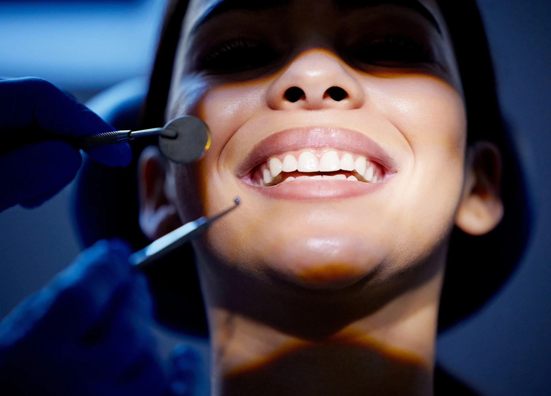 Close-up of a woman's bright smile lit in blue as a dentist holds a mirror and explorer