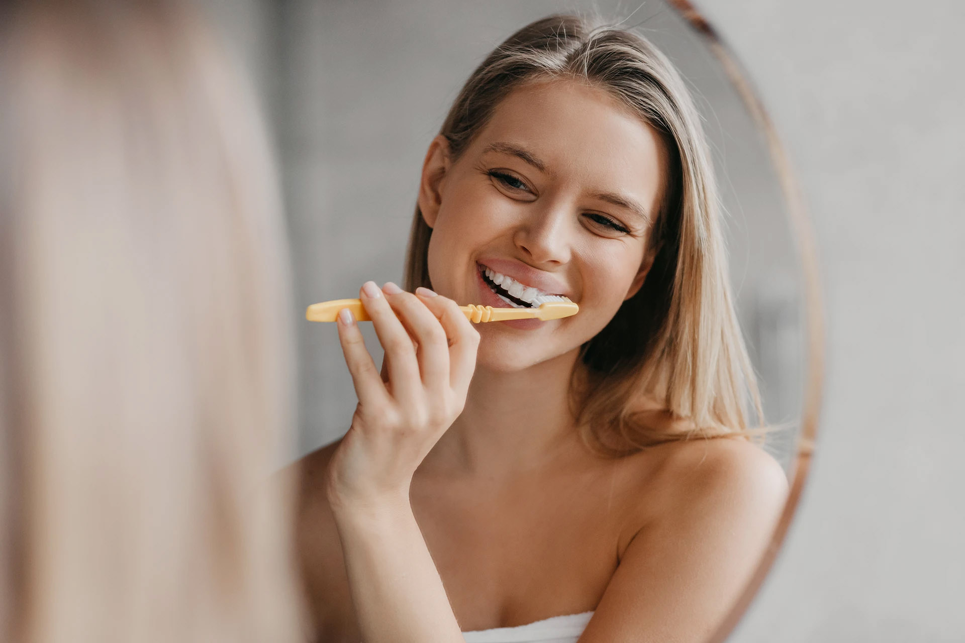 Smiling blonde woman brushing her teeth with a yellow toothbrush while looking in a mirror