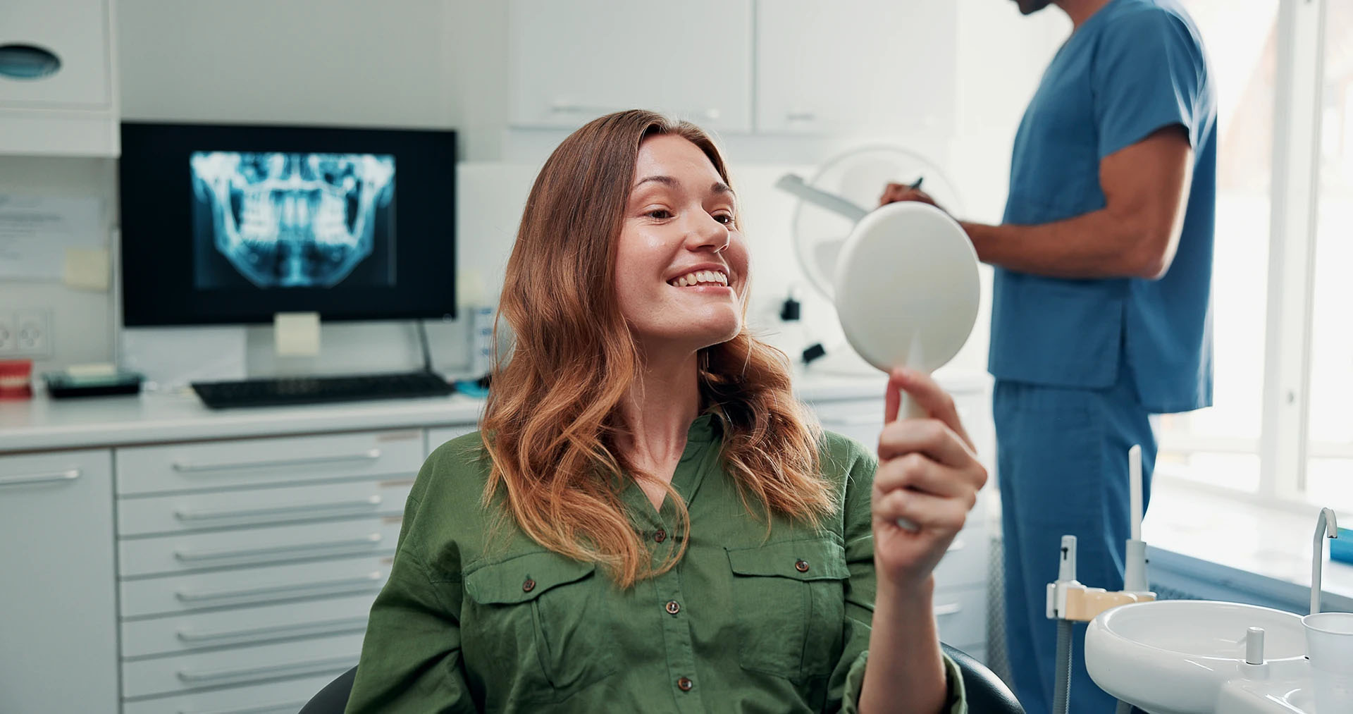 Smiling woman in a dental chair looking at her teeth in a hand mirror with the dentist working behind her