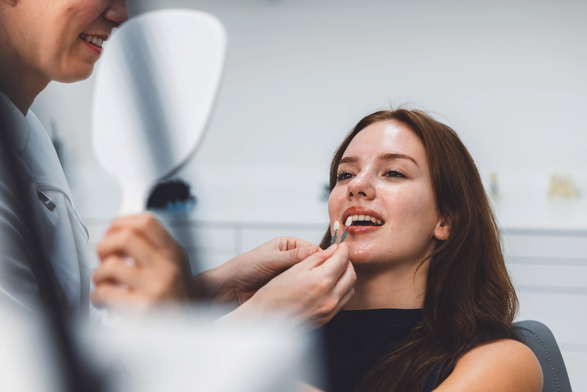 Smiling woman looking in a hand mirror as a dentist compares a veneer shade sample to her teeth