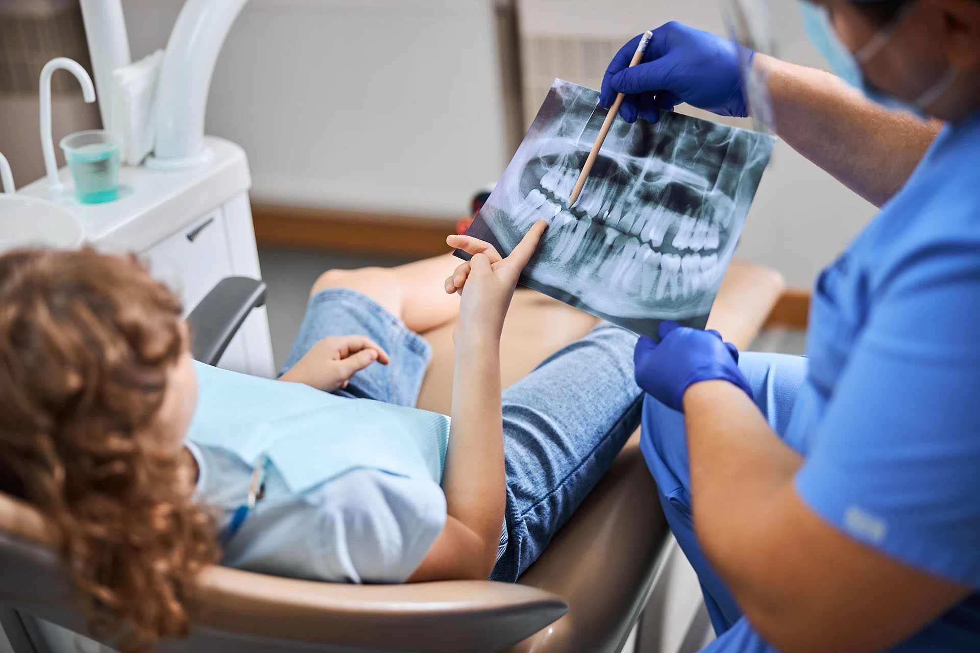 Dentist in blue scrubs showing a panoramic X-ray film to a young girl in the dental chair