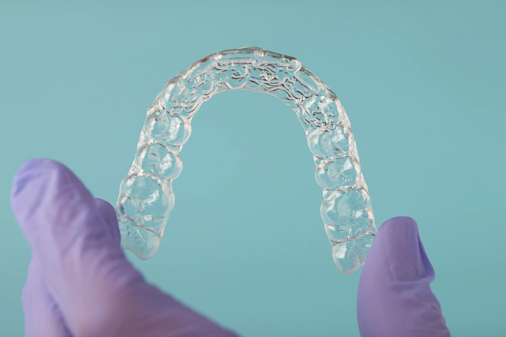 Purple-gloved hands holding a transparent clear aligner against a teal background
