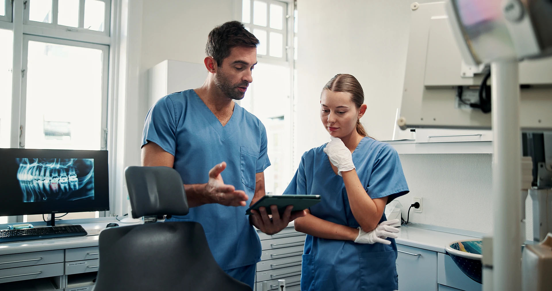 Two dental professionals in blue scrubs discussing a case over a tablet in the operatory