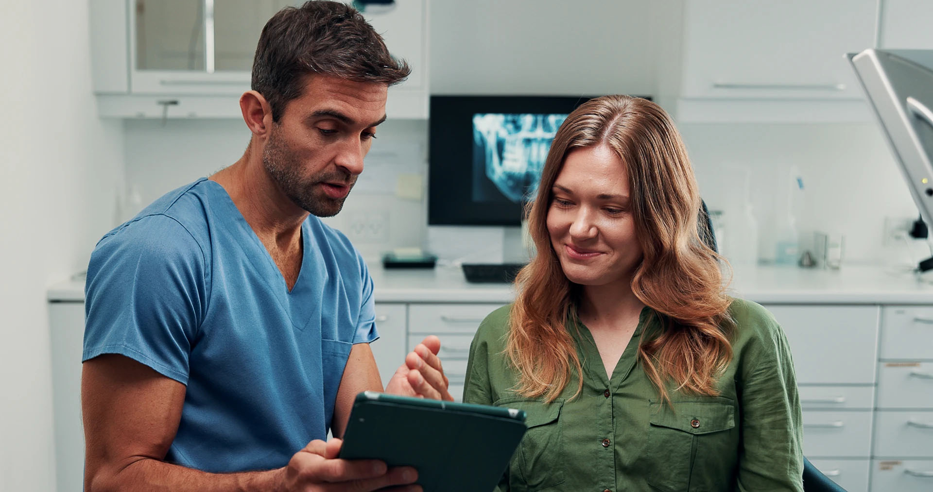 Dentist in blue scrubs discussing treatment on a tablet with a woman patient, an X-ray on the wall behind