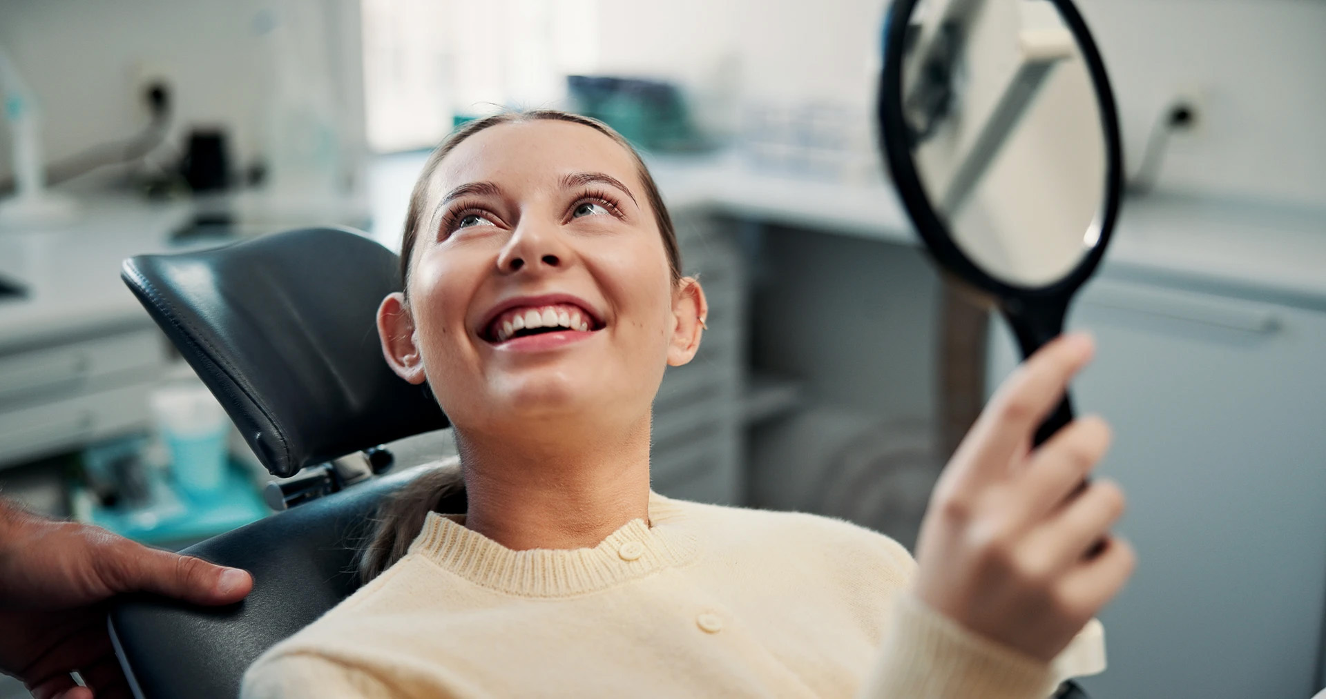 Young woman in a dental chair smiling with delight as she looks at her teeth in a hand mirror