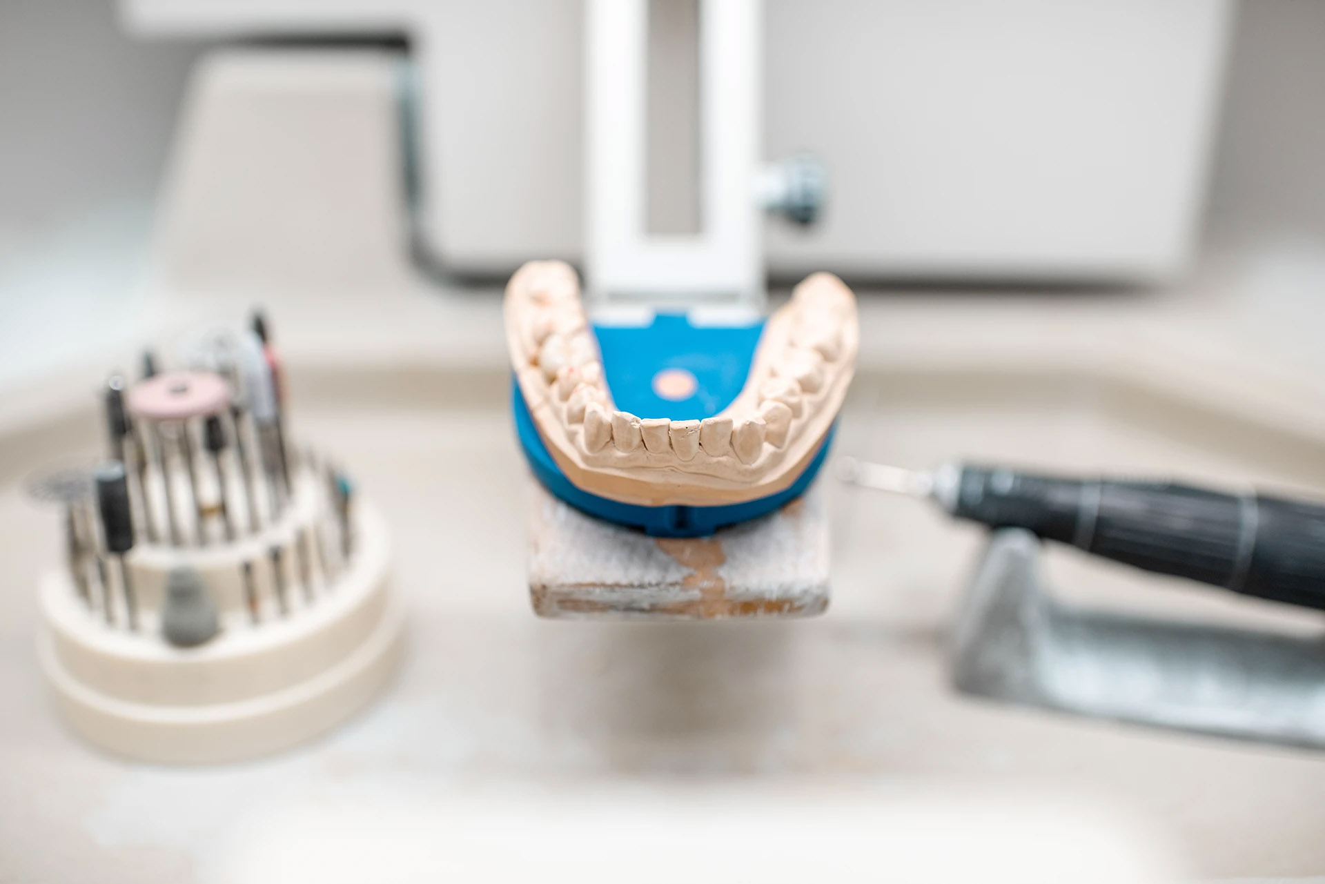 Plaster dental model on a blue articulator beside polishing burs and a handpiece on a lab bench
