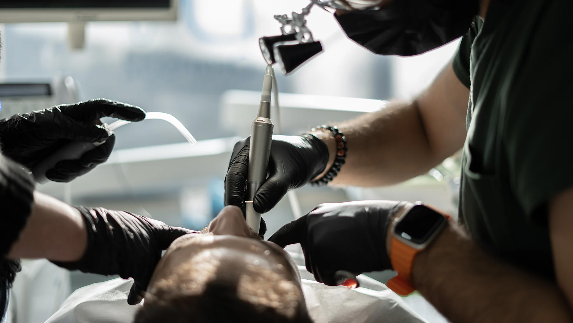 Two masked dental professionals in black gloves working on a patient with a handpiece and suction