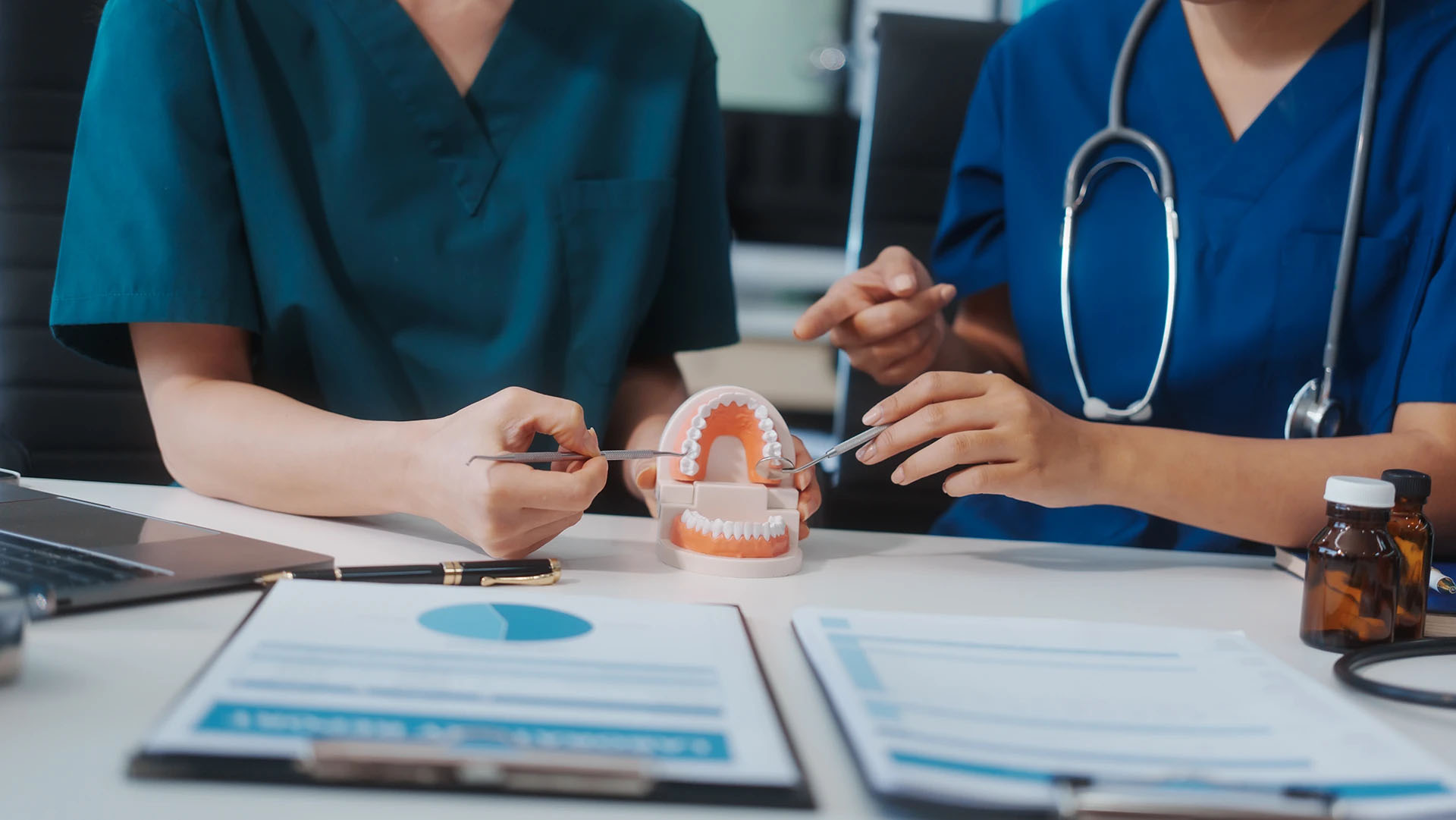 Two dental professionals in scrubs examining a teeth model with instruments at a desk
