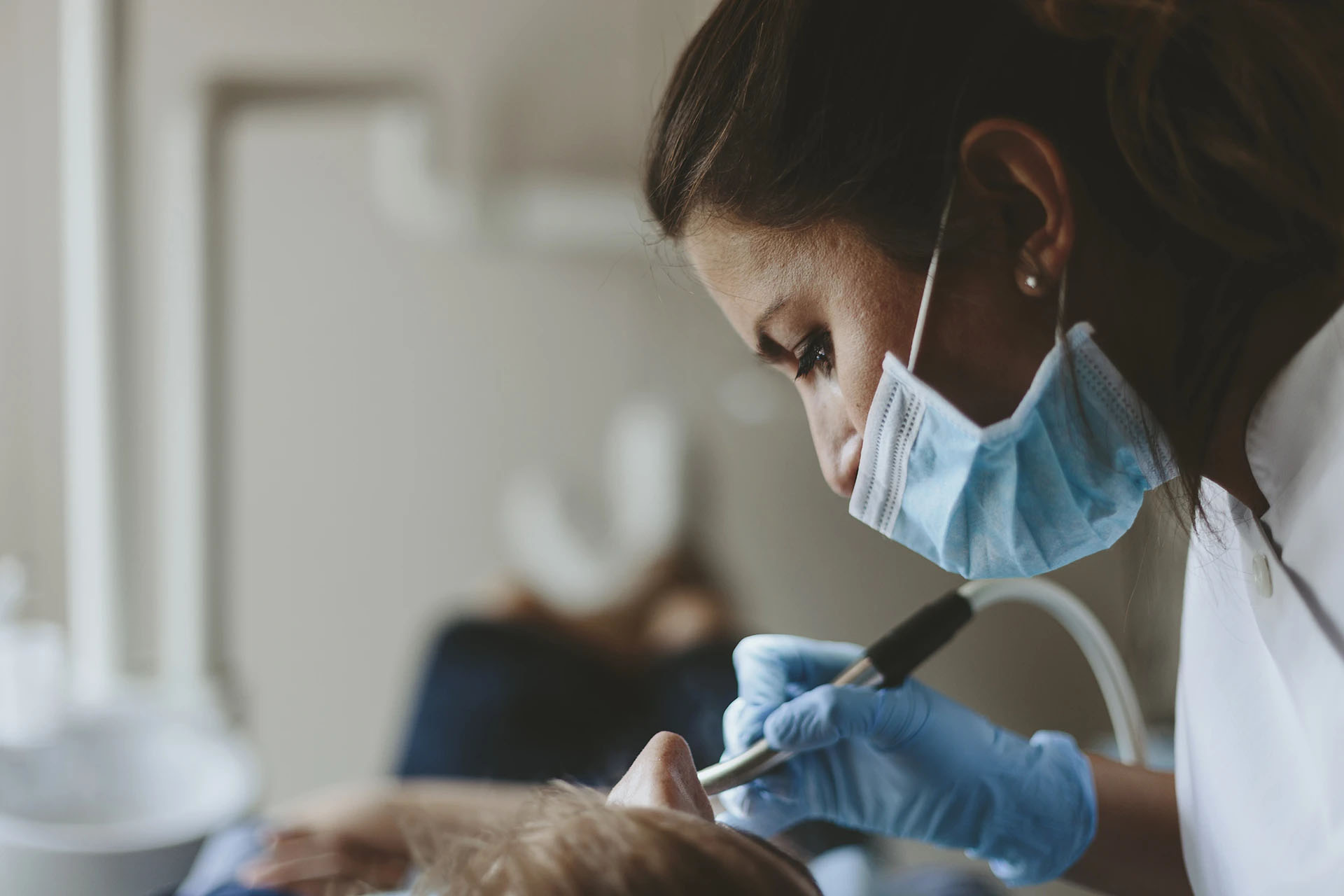 Masked female dentist in blue gloves leaning over a patient using a suction tool in soft light
