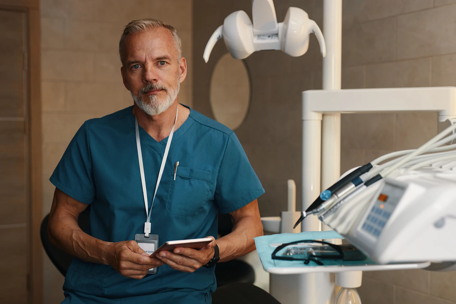 Mature male dentist in teal scrubs holding a tablet in a dental treatment room