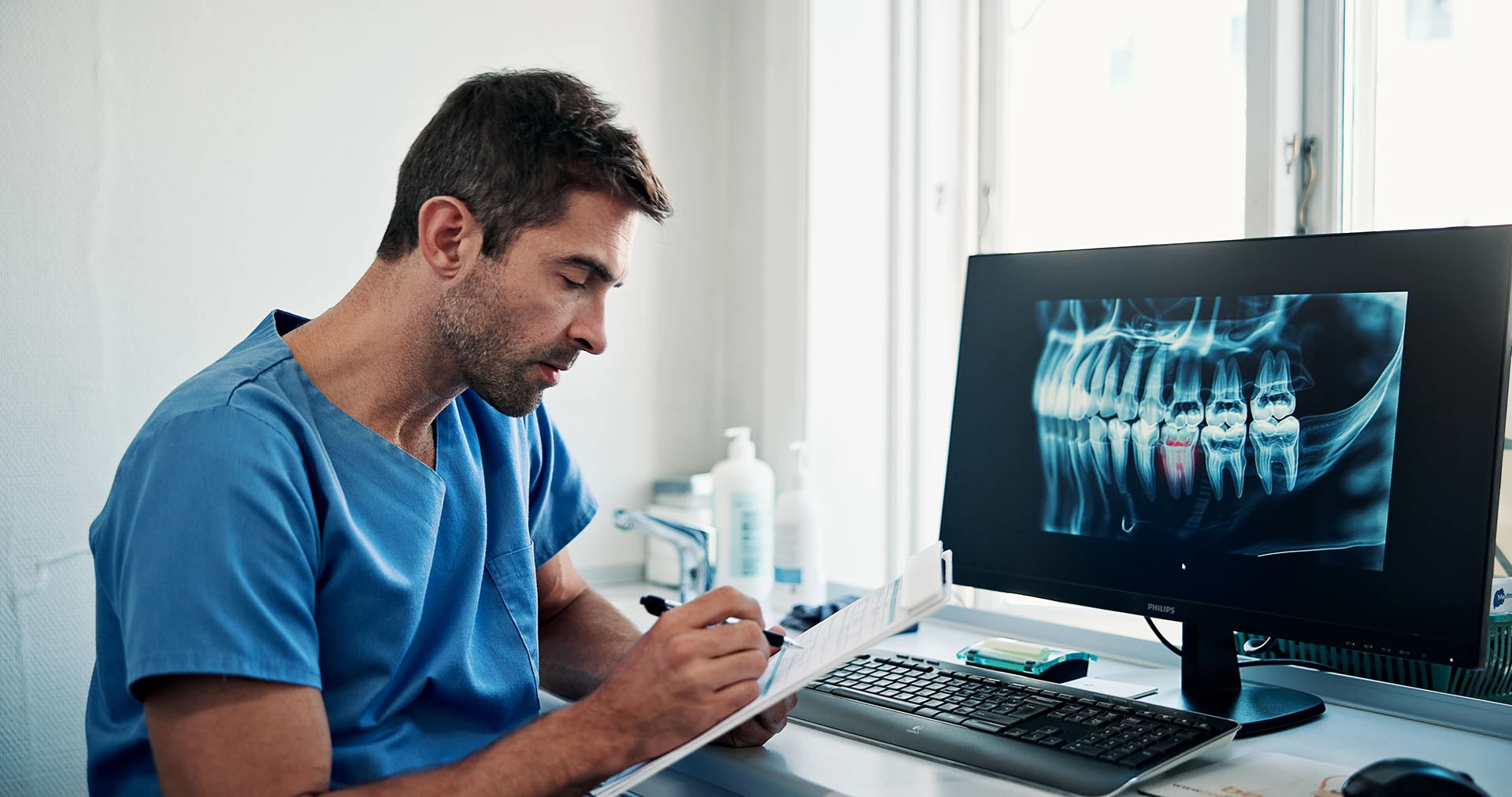 Male dentist in blue scrubs writing on a chart at his desk beside a monitor showing a dental X-ray