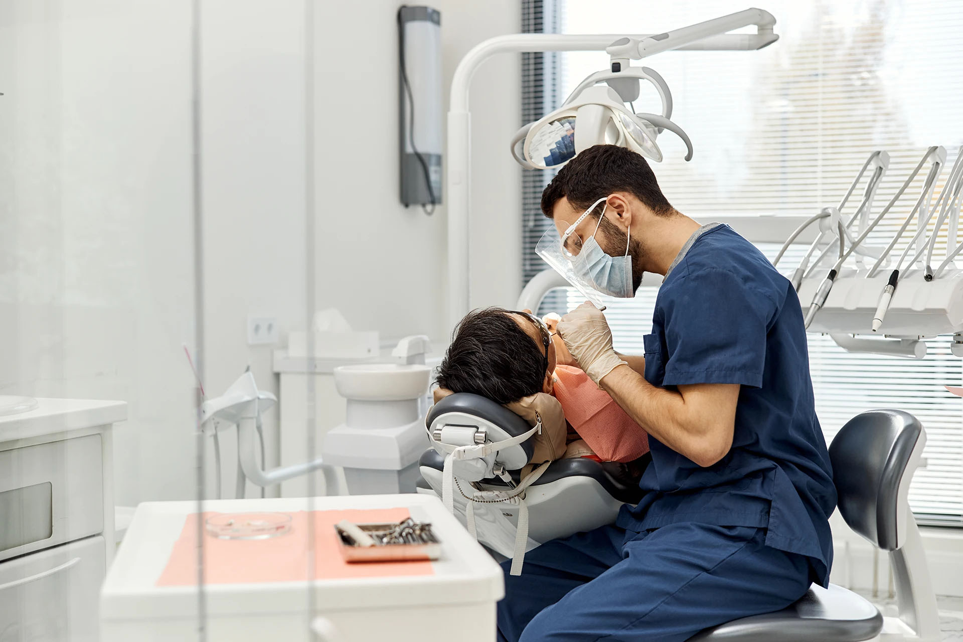 Dentist in blue scrubs and face shield leaning over a reclined patient during a procedure