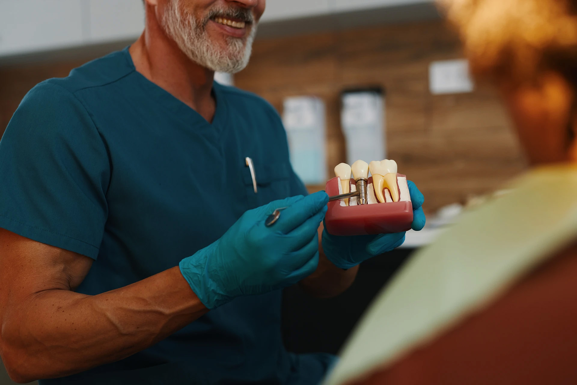 Mature dentist in teal scrubs pointing to a dental implant demonstration model for a patient