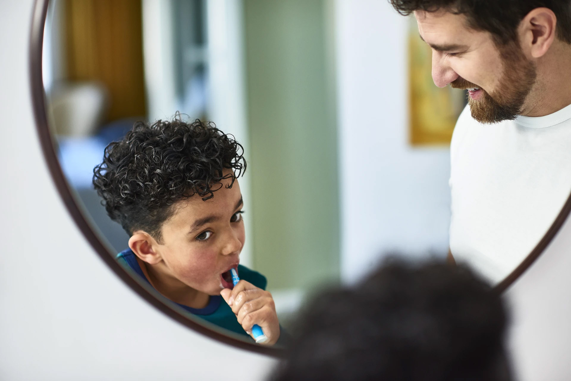 Young boy brushing his teeth at a bathroom mirror while his smiling father looks on