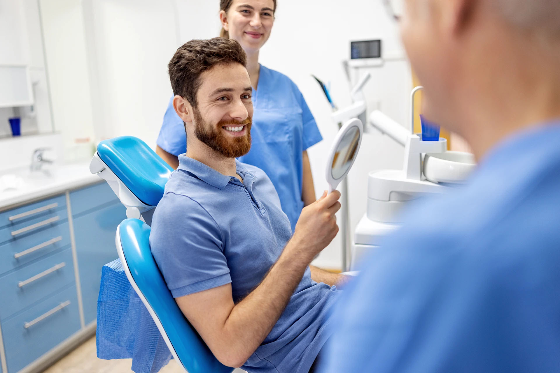 Smiling bearded man in a dental chair holding a hand mirror with two dental staff nearby