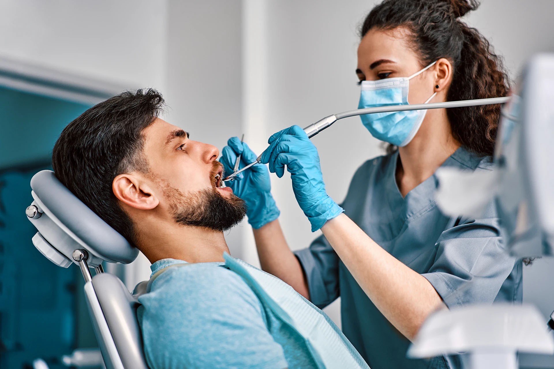 Bearded man reclined in a dental chair as a masked dentist works in his mouth with a suction tool