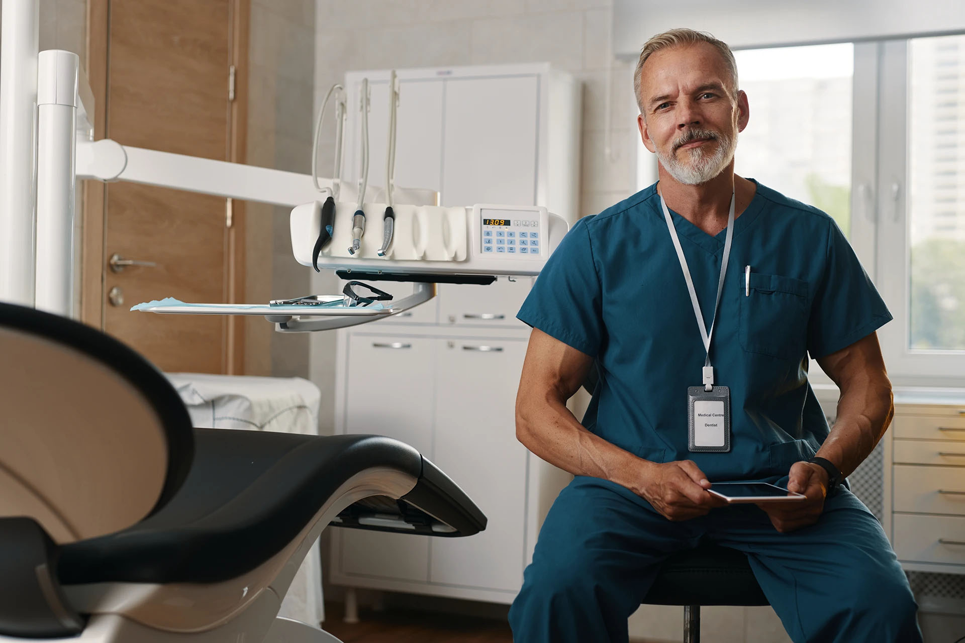 Mature male dentist in teal scrubs with a lanyard holding a tablet beside the dental chair