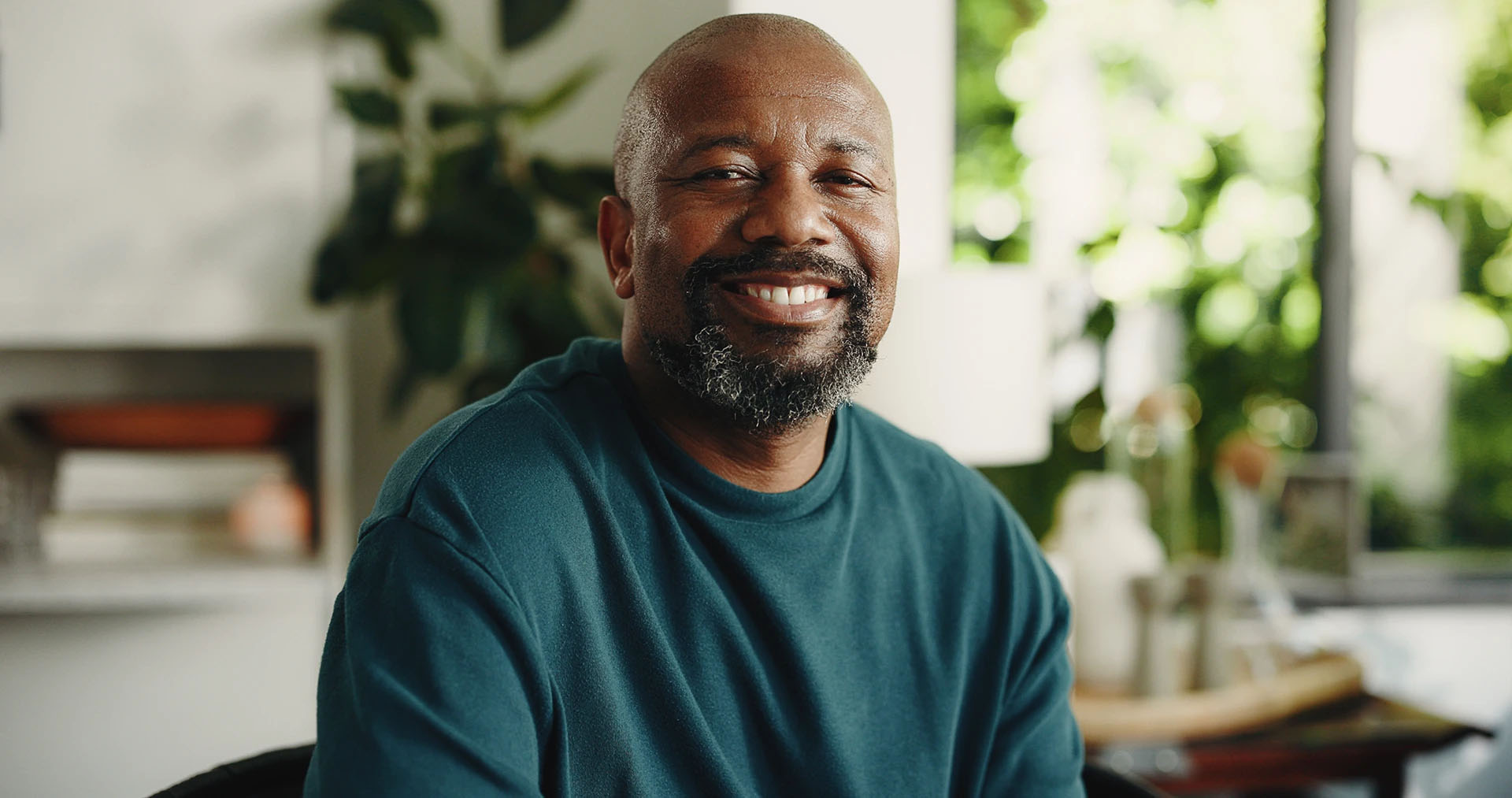 Smiling mature Black man with a gray beard in a teal sweater at home with plants behind him