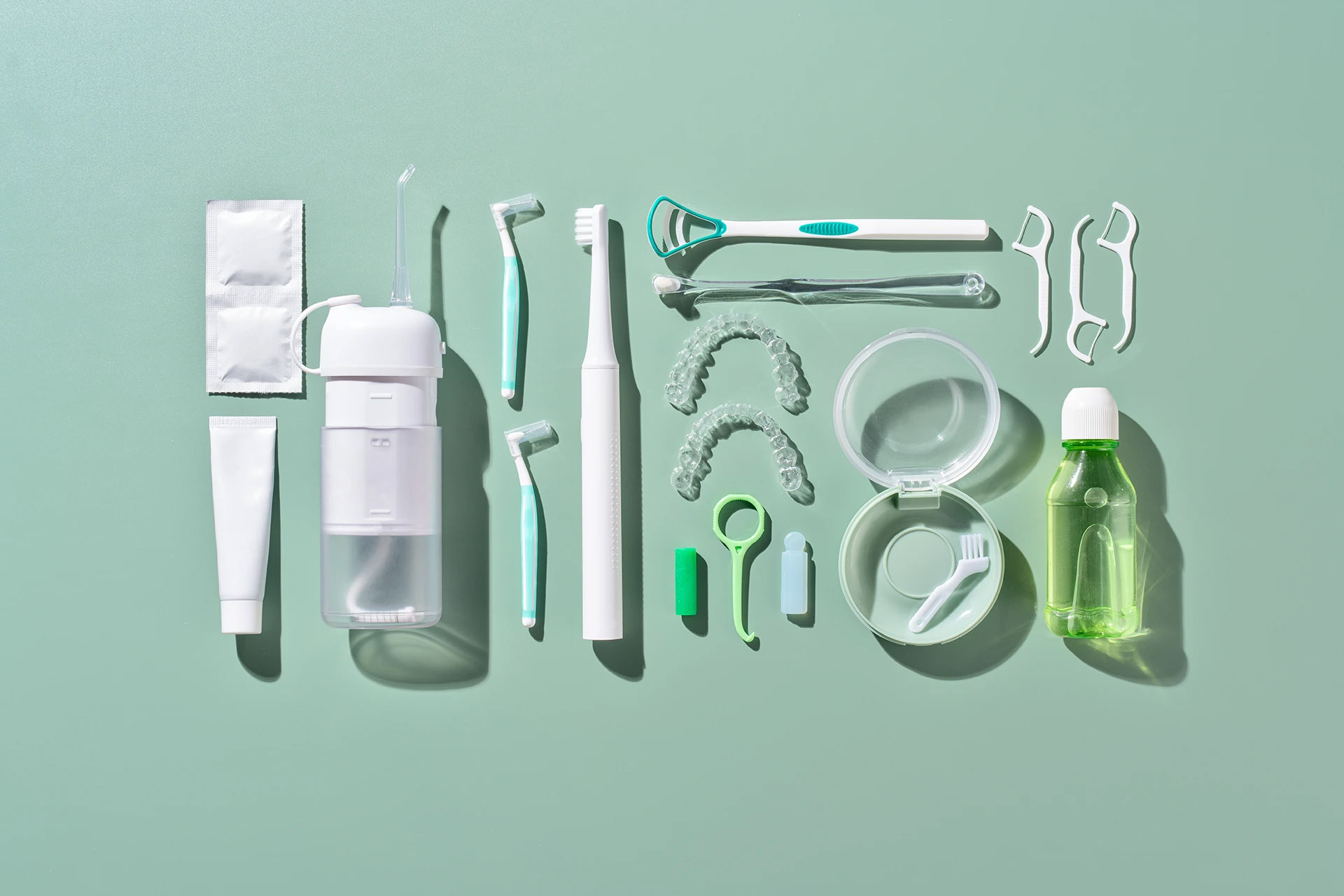 Flat lay of oral hygiene products including toothbrush, floss, tongue scraper, clear aligners and mouthwash on a mint background