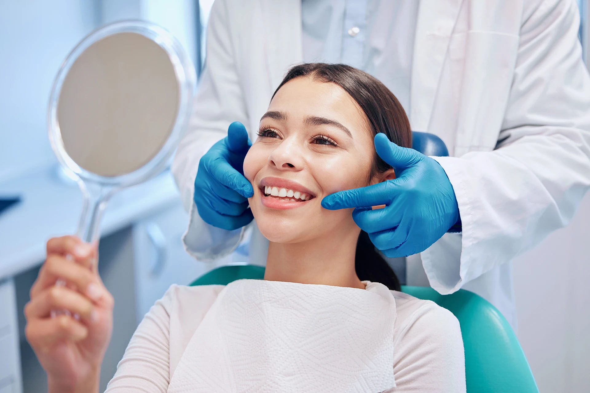Smiling young woman in a dental chair looking in a hand mirror as a dentist in blue gloves frames her face