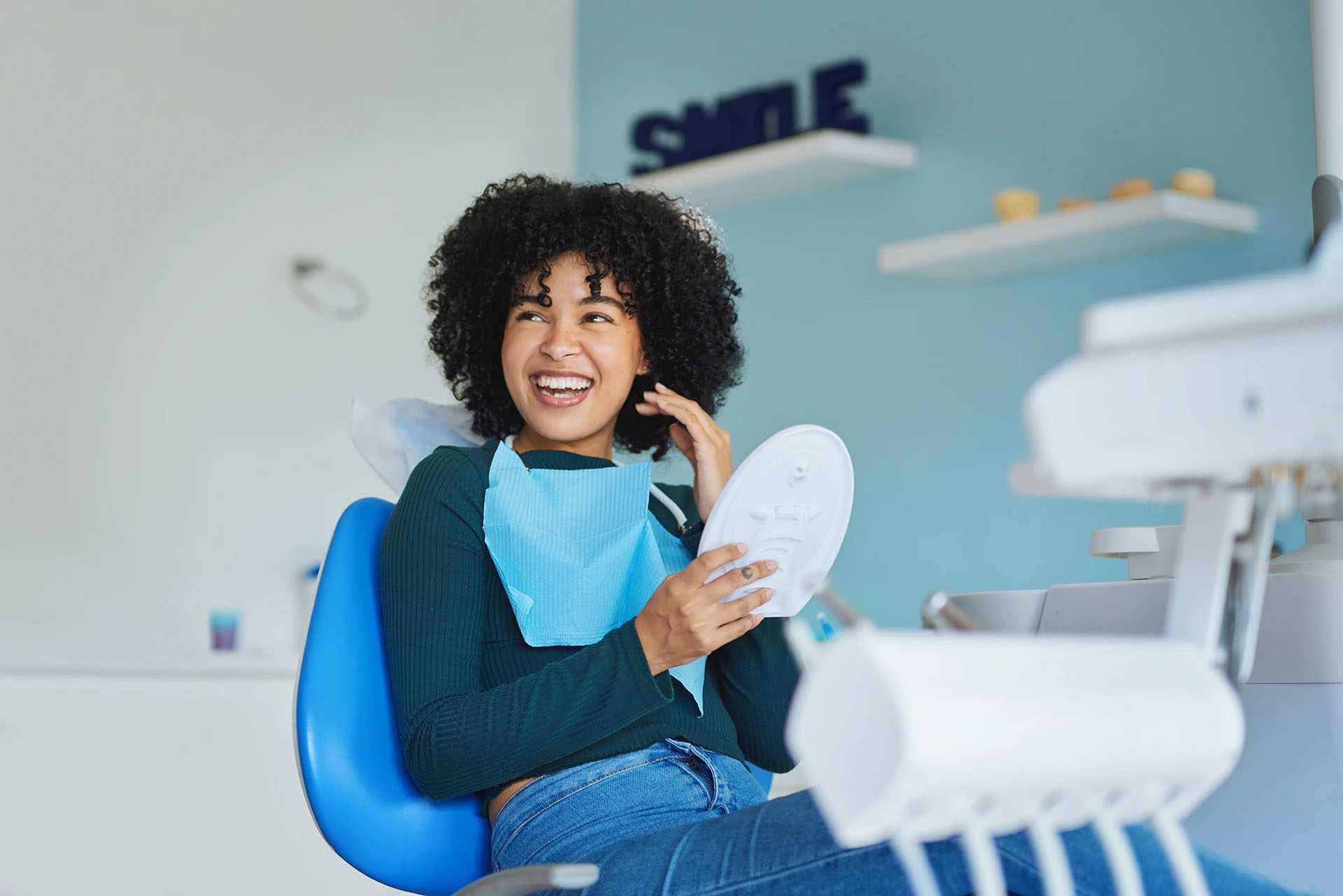 Young woman laughing in a dental chair holding a hand mirror with a SMILE sign on the wall behind her