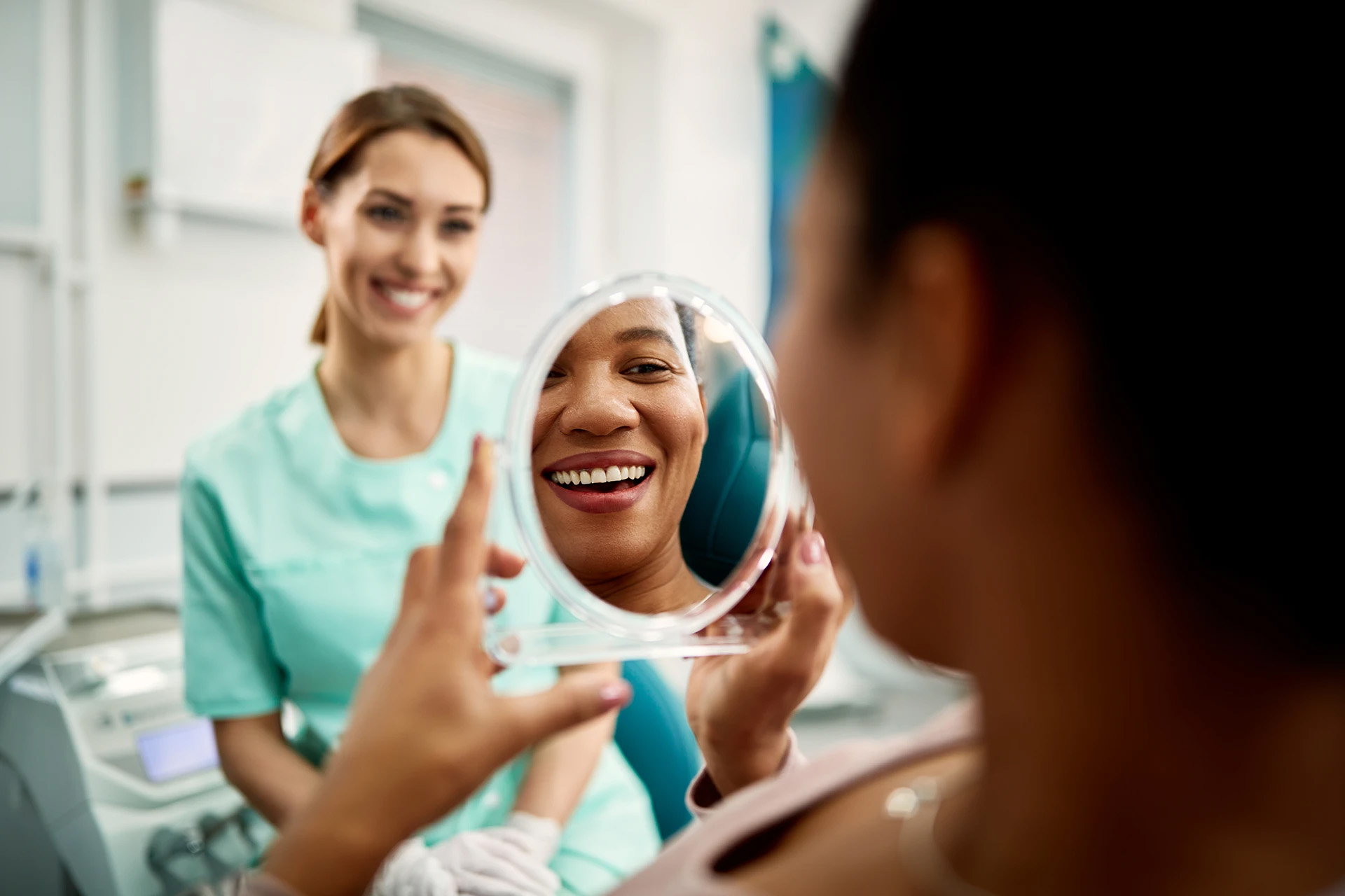 Woman in a dental chair admiring her smile in a hand mirror while a smiling hygienist looks on
