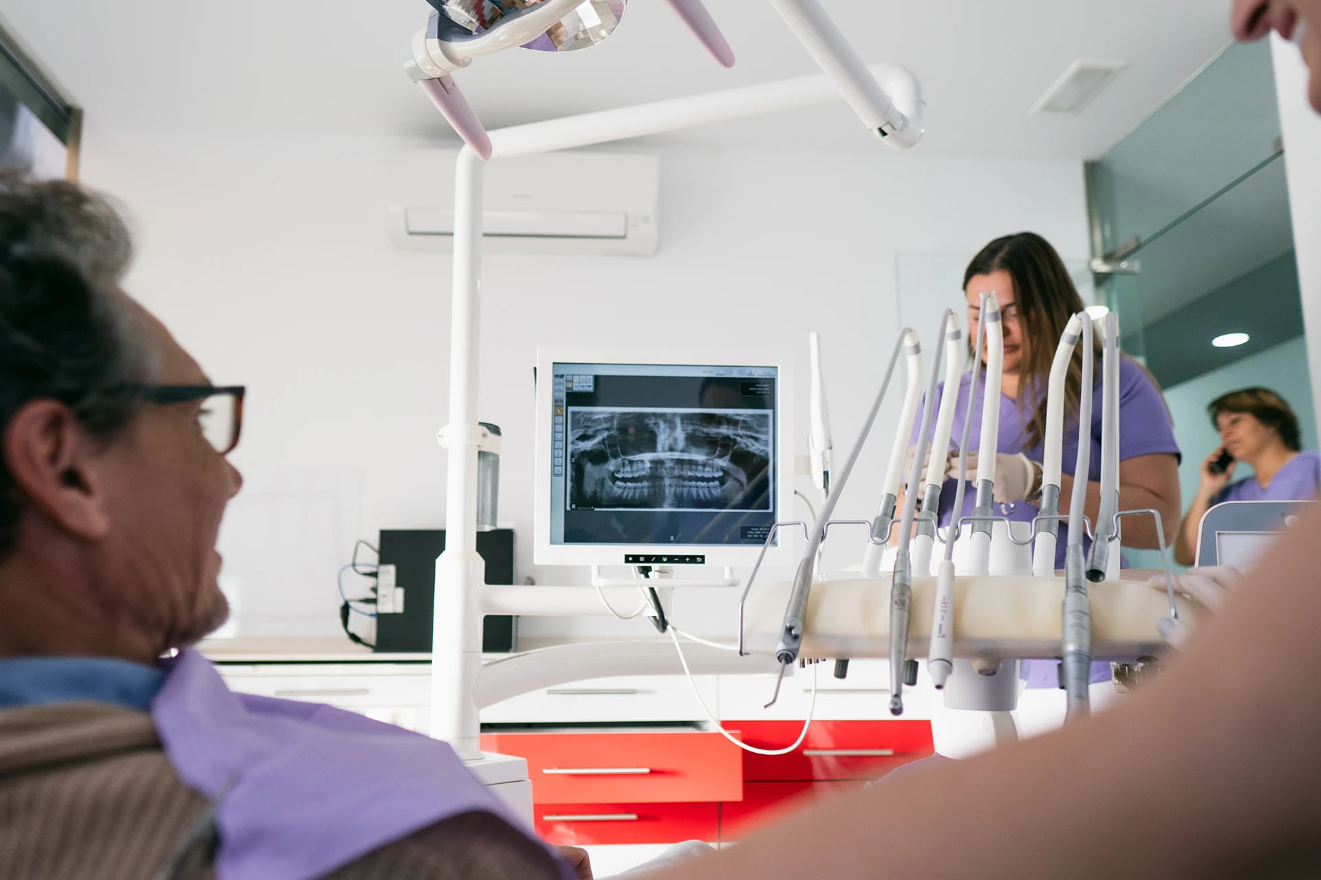 Older man in a dental chair looking at his panoramic X-ray on a monitor while staff work in the background