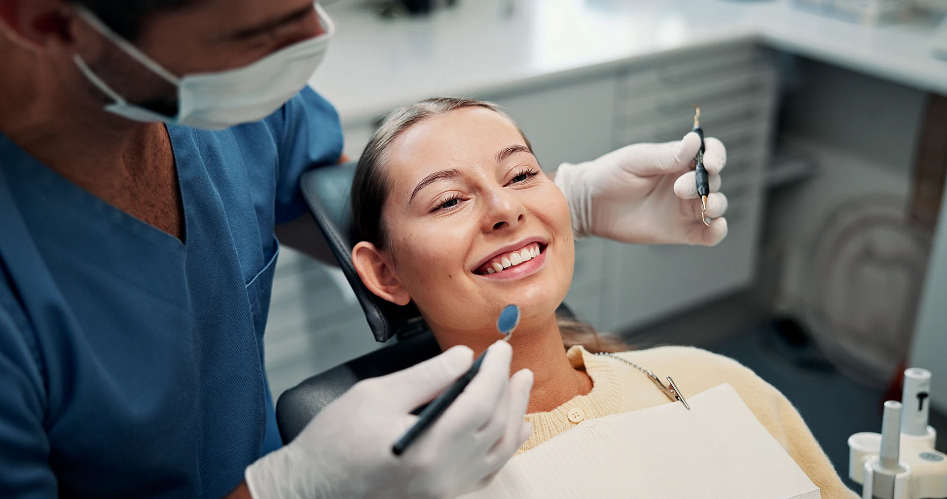 Masked dentist in blue scrubs holding a mirror and explorer beside a smiling woman patient
