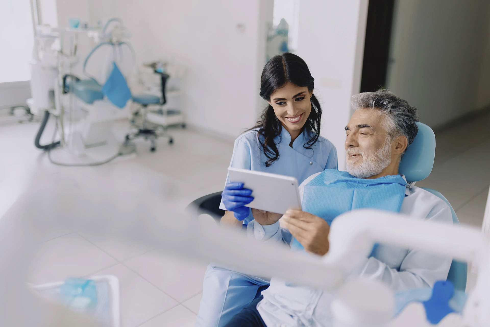Smiling dentist in blue showing a tablet to a senior man in the dental chair