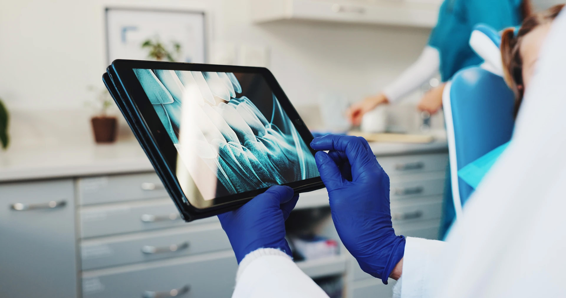 Dentist in blue gloves holding a tablet showing a dental X-ray with a patient reclined in the background