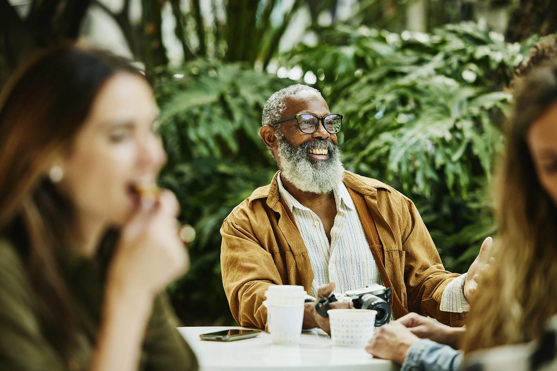 Older man with a gray beard laughing at a cafe table with friends among green plants
