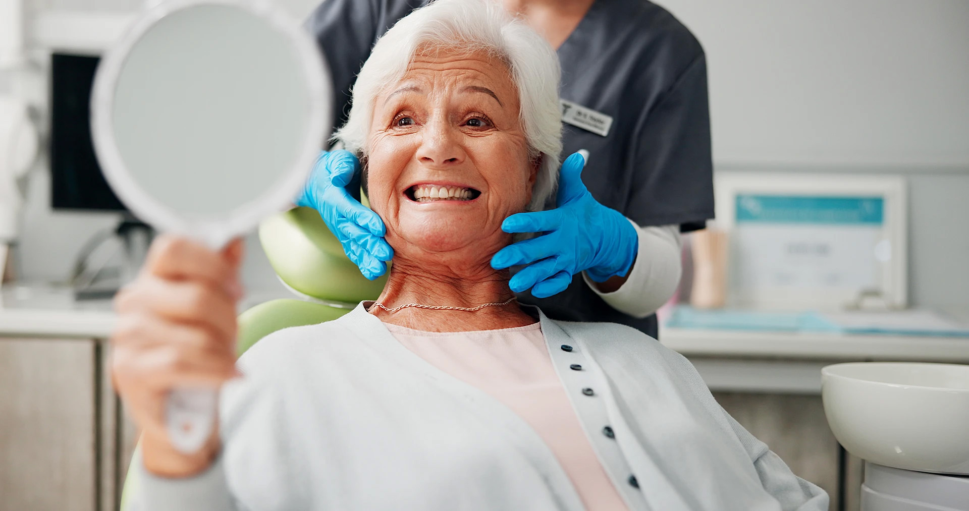 Happy senior woman looking in a hand mirror as a dentist in blue gloves frames her face
