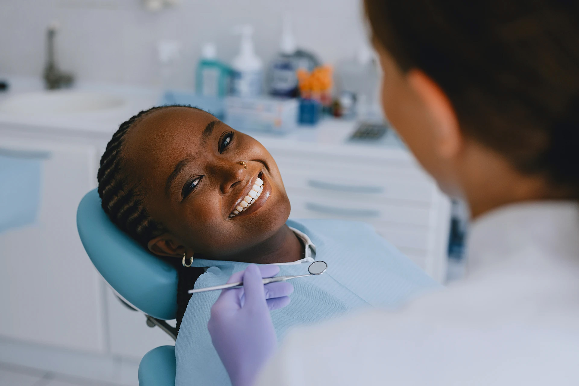 Black woman with braids smiling in a blue dental chair as a dentist in purple gloves prepares to examine her