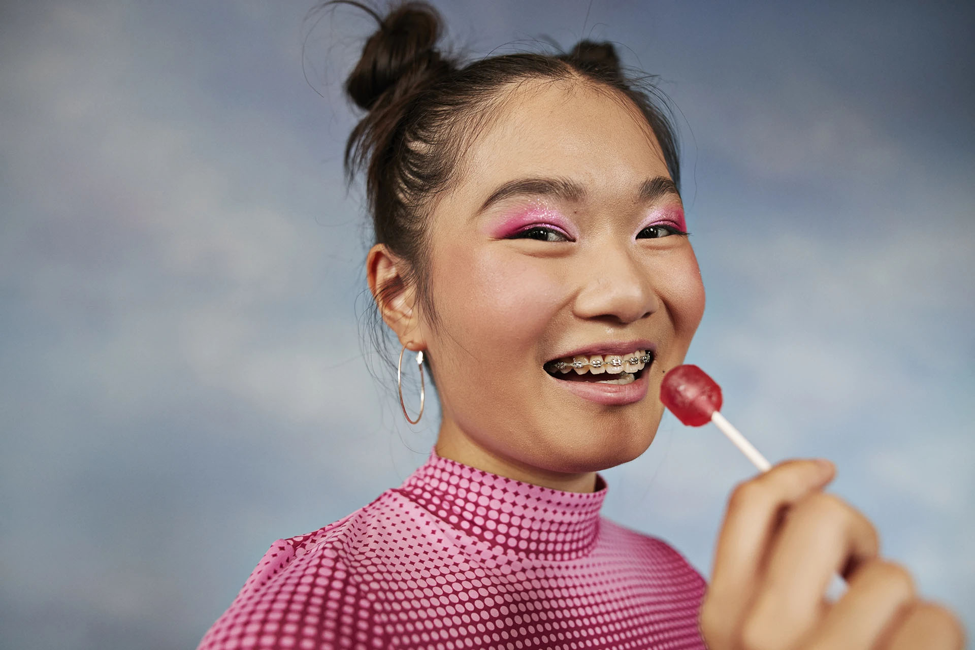 Smiling teenage girl with metal braces holding a red lollipop against a blue sky backdrop