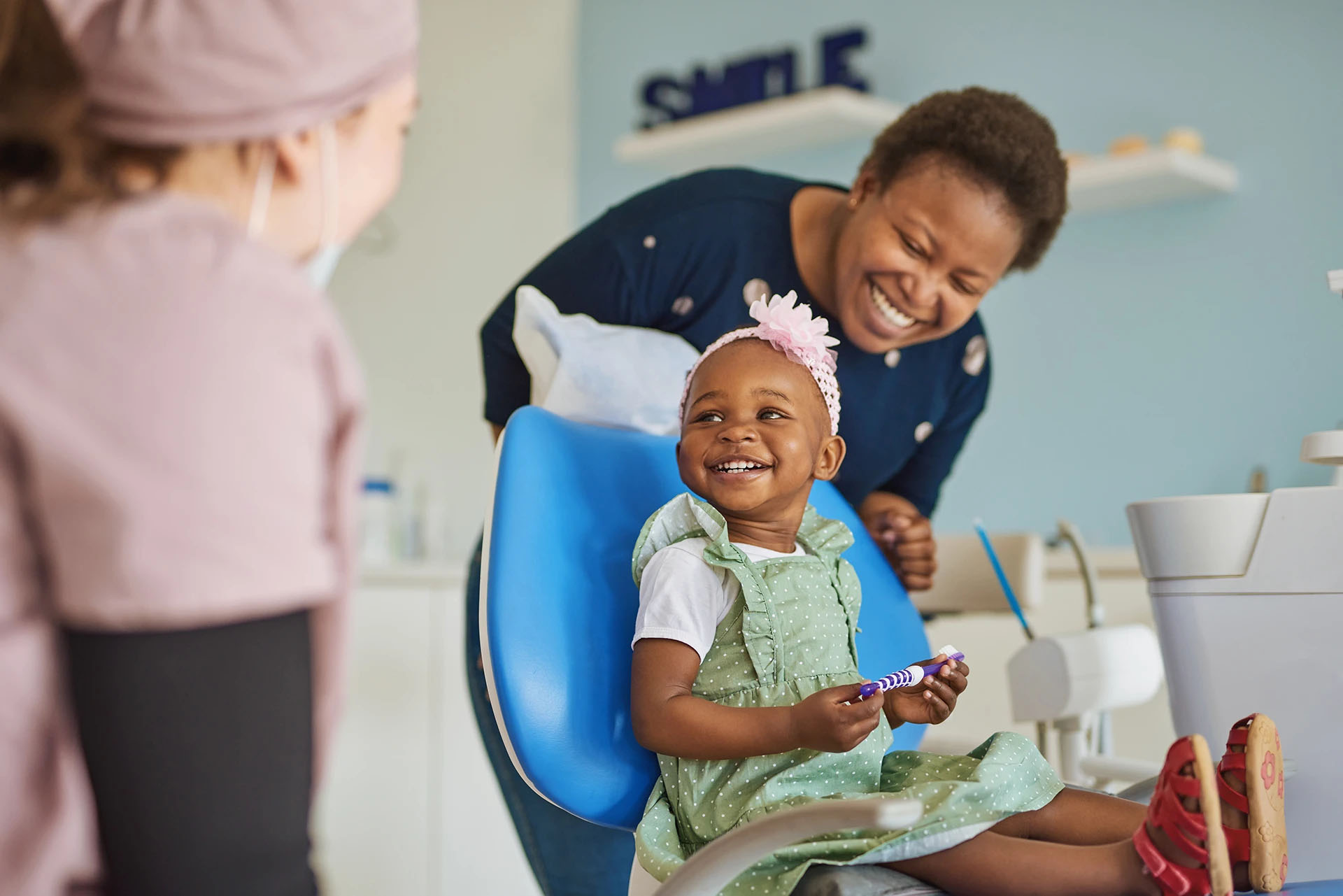 Smiling toddler in a green dress holding a toothbrush in a dental chair with two friendly dental staff