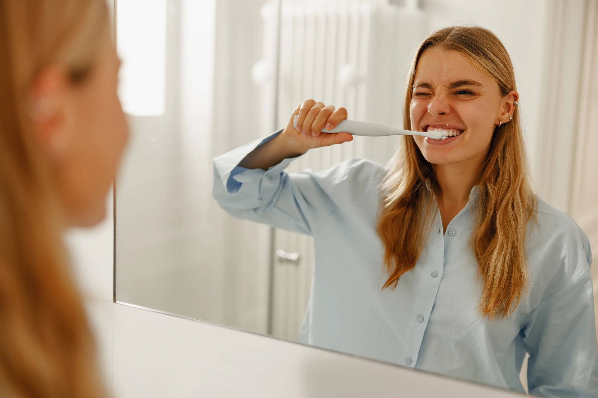 Blonde woman in a light blue shirt brushing her teeth and winking in the bathroom mirror