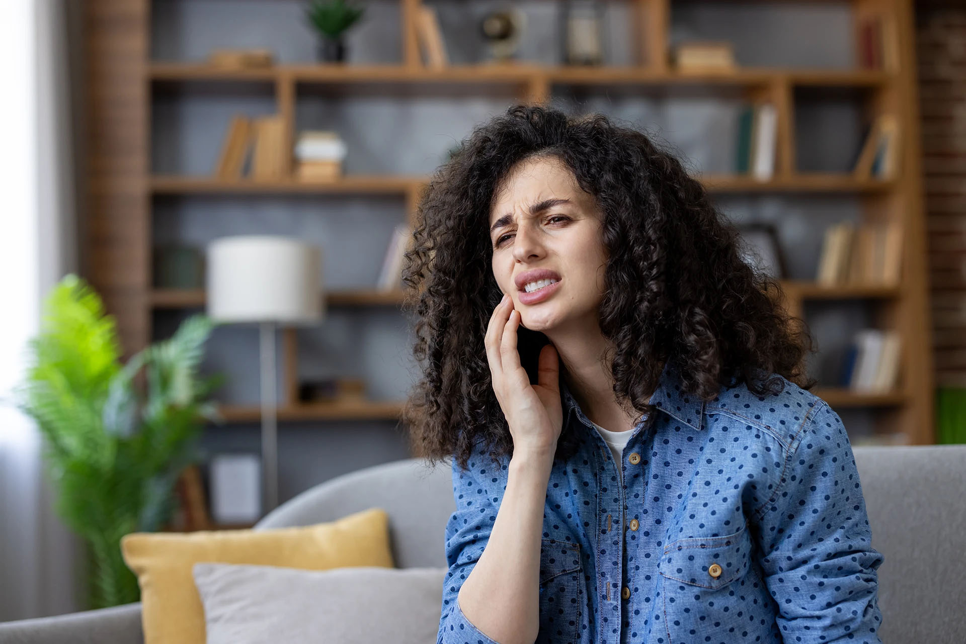 Woman with curly hair wincing and holding her cheek from tooth pain at home