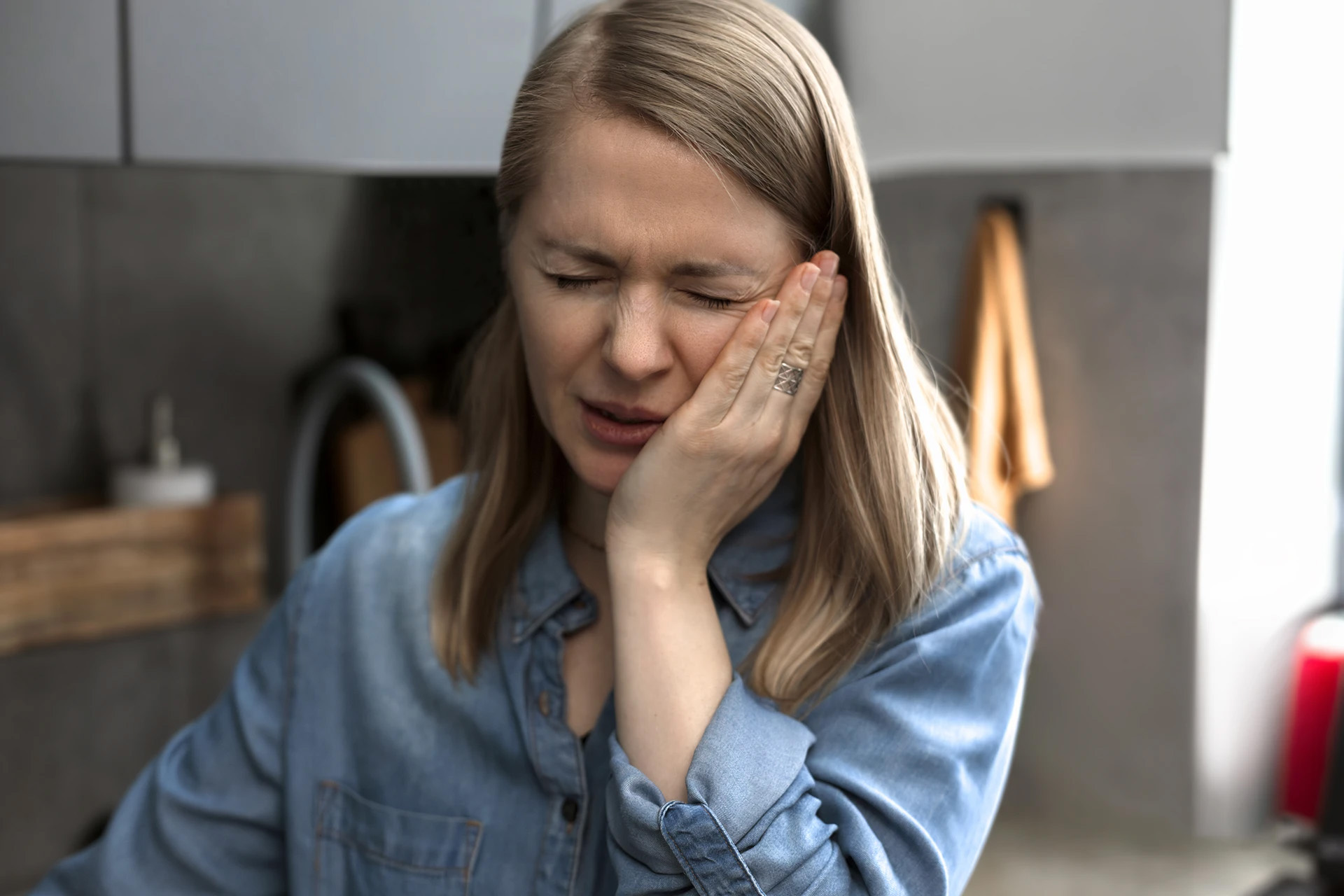 Woman in a denim shirt wincing and holding her cheek in pain from a toothache