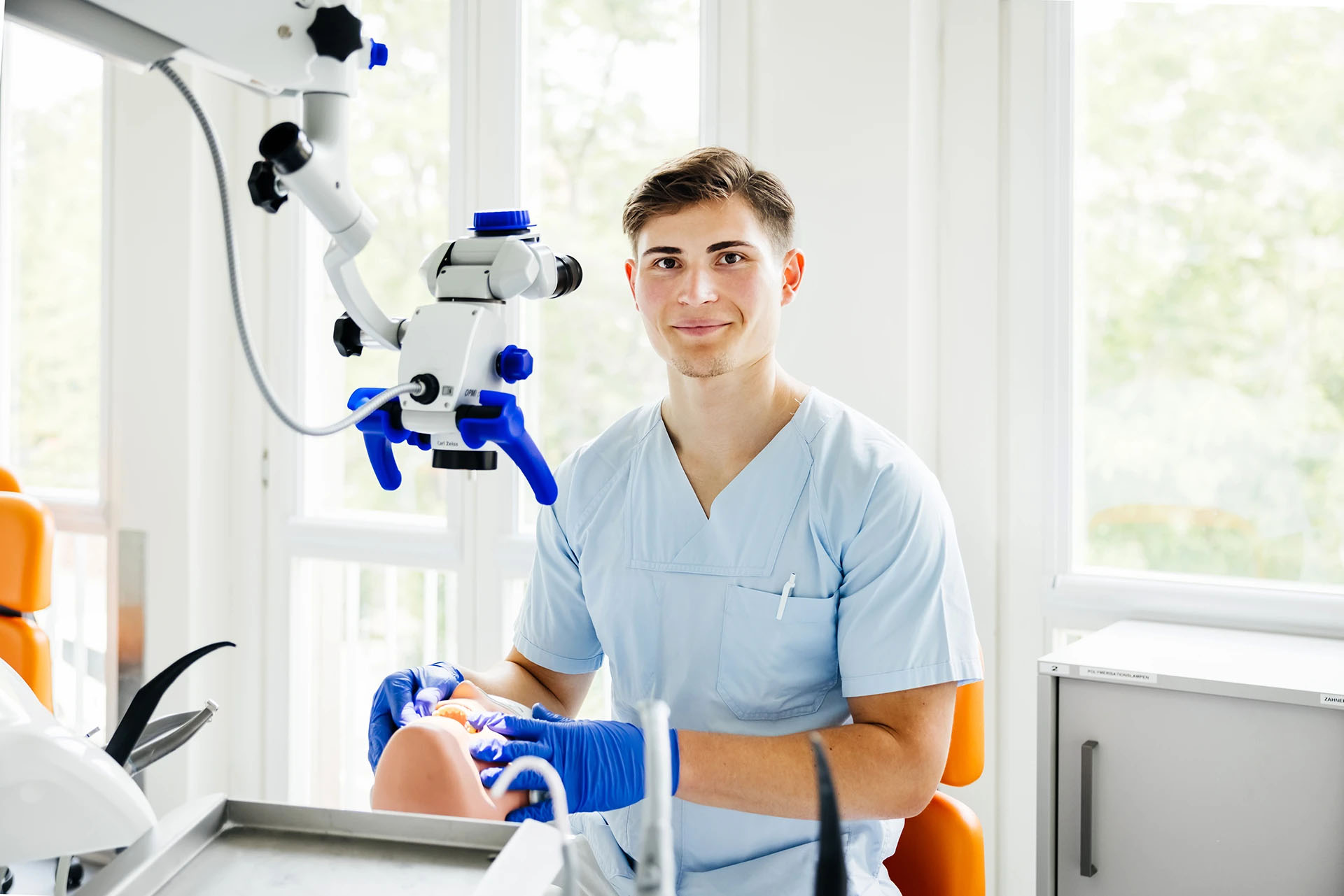 Young male dentist in blue scrubs smiling beside a dental operating microscope in a bright office