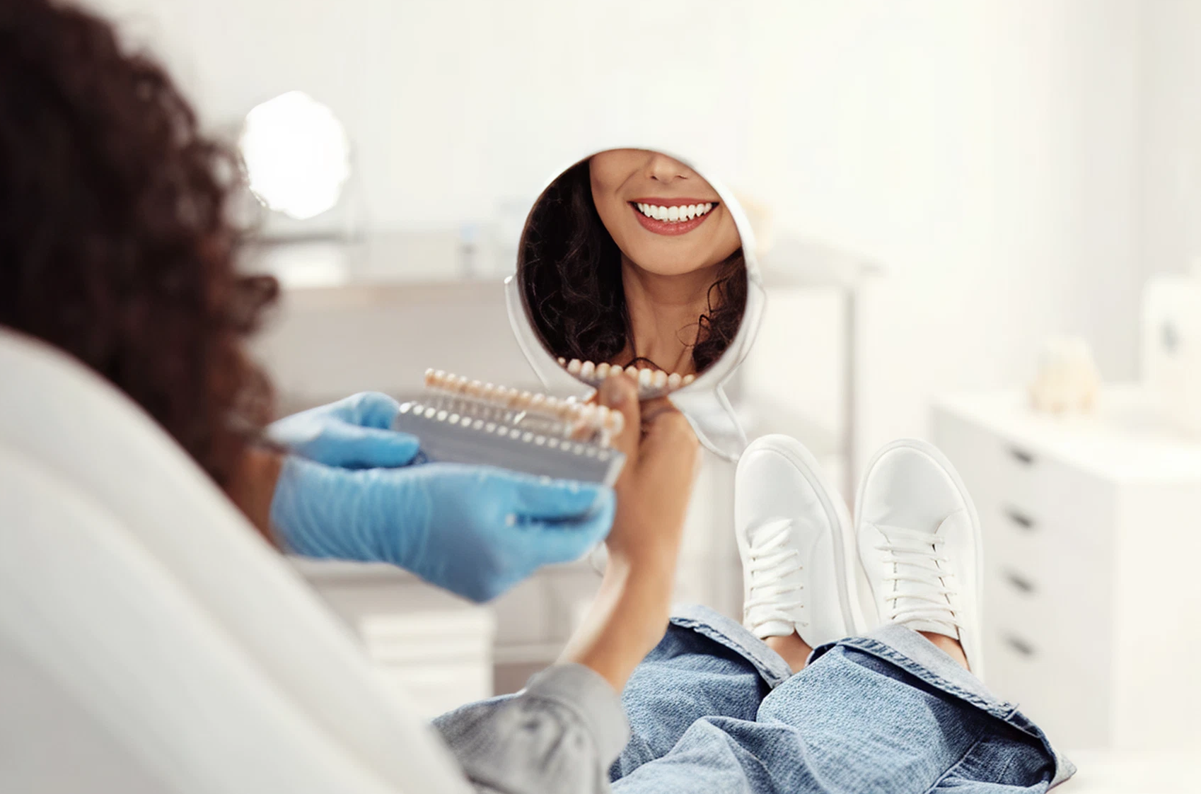 Woman in a dental chair smiling into a hand mirror as a dentist holds a shade guide